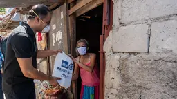 A man wearing a mask hands a bag to an older woman, who is standing in the doorway of a house.