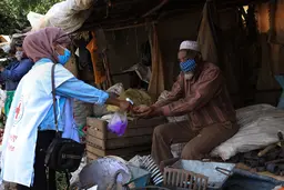 Young woman wearing a red cross vest sprays an old man hands. Both wear masks. 
