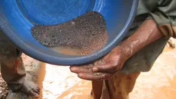 Close-up of a bucket containing soil/water/small rocks being poured into a waiting, outstretched hand