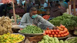A man wearing a mask is sitting surrounded by vegetables: green beans, tomatoes, spring onions.