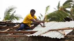 A man sits awkwardly using a hammer to try and fix corrugated roofing to a house.