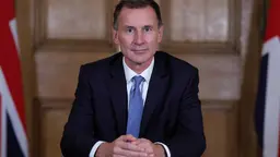 A man in a suit sits at a table with his hands crossed in front of him.