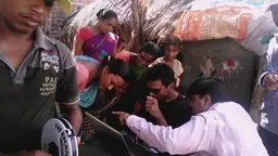 A group of people gather around a man seated with a laptop computer and look and point at the screen