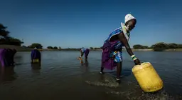A woman in the foreground bends down to gather water in a yellow barrel. More women can be seen doing the same in the background.