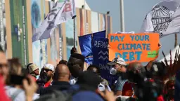 Mass of people walking with flags and posters. A sign reads "pay your climate debt"