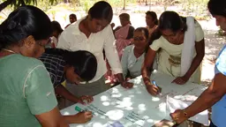 A group of women stand around a table and write on a white piece of paper.