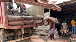 Man carrying a bag over his next next to a truck full of bags.