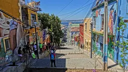 Street with colourful buildings and people standing in the shade. A body of water can be seen in the background.