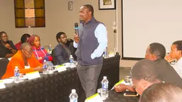 Man stands holding a microphone surrounded by seated people.