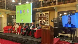 A seated panel of four people listen to a speech given by a remote colleague on a television screen positioned to their left.