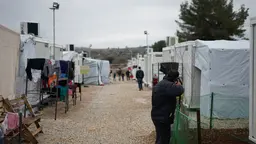 Man in black jacket walking among temporary tents and shelters.