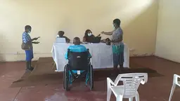 A woman wearing a mask receives money from another mask-wearing women behind a desk. Three other people in the bare room (one in a wheelchair) are standing/seated far apart.