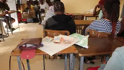 Papers are strewn on an empty desk as people sit at desks in a room.