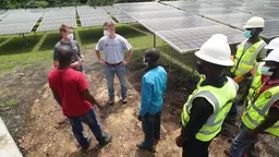 A man wearing a mask, standing in front of a large number of solar panels, talks to a wider group of people who are mostly wearing high visibility clothing and hard hats. 