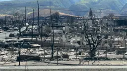 A mess of burned out buildings and vehicles in front of imposing mountains in the background.