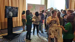 As a group people mill around a room and talk with one another at an event, an elevated TV screen shows the transcript of speakers at the event.