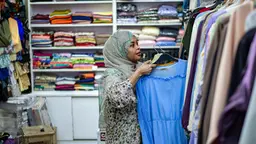 A woman hangs up clothes on a rail in a shop, with clothes on shelves behind her.