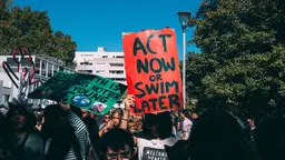 A woman on a protest march holds a sign above her head, saying: 'Act now or swim later'. The sign has a vivid red background.