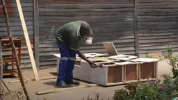 A man, weariug a mask, bends over a piece of furniture he is making outside his house. 