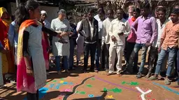 A woman, surrounded by a standing group of people outside, points to the floor where there are brightly coloured markings.