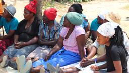 A group of women sit on the floor outside listening to a discussion.