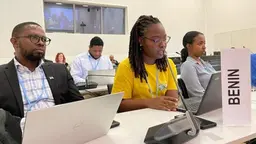 Woman sitting at a table speaks to a microphone. A sign saying "Benin" stands in front of her.