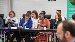 Five woman sitting behind tables, listening and taking notes.