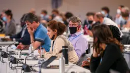  People at tables with microphones and laptops, a woman is speaking.