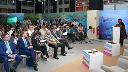A large group of seated people listen to a women speaking at a podium.