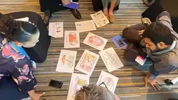 People sit on the floor around a series of printed cartoons and illustrations in the centre.
