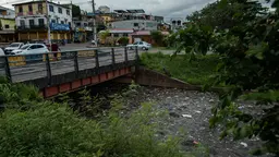 Garbage collects in a stream under a bridge. In the background, cars are parked in front of shops and homes.