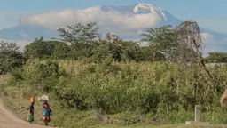 Two women walk along a road beside green undergrowth and fields, with a mountain in the background entering the clouds.