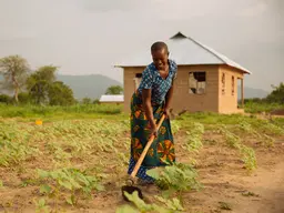 A woman farmer tends her crops with a hoe in front of a small house.