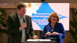 A man in a suit holding a microphone laughs along with a woman standing next to him behind a podium. 