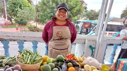 A woman stands behind a market stall of fresh vegetables, smiling proudly.