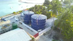 Two large, round water tanks. There is sea in the background, and palm trees around.