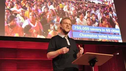 A bearded man stands at a podium with a screen behind him.