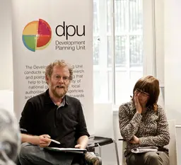 A seated man talks to a group, while a women alongside watches on. Behind them is a board advertising DPU - the Development Planning Unit at UCL.