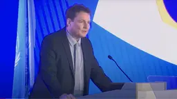 A man in a suit leads forward on a podium making a speech.