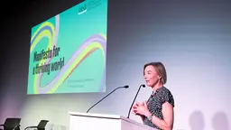 A woman stands at a podium giving a speech, while in the background is an image of a publication cover, saying: "Manifesto for a thriving world".