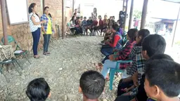 Two people stand in an outside area, surrounded by seated youngsters in a semi-circle.