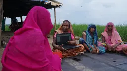 Women sitting down in a circle. One of them is holding a laptop and talking.