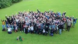 A large group of people stand in a green space and look up waving at a camera. 