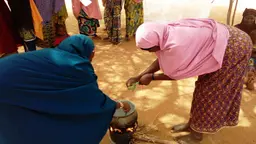 Two people bending down, cooking in a pot set over an open fire. This was part of a cooking demonstration for the village.