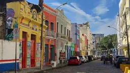 Colourful murals adorn similarly bright-coloured buildings alongside a street, under a blue sky,