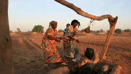 With a dusty backdrop, two women grab rope leading over a bar to a bucket, as they struggle to get water out of a well.