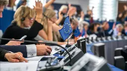 Representatives in the European Parliament voting on an issue. Some have their hands raise while others are writing in notebooks.