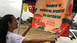 Person holding gup and reading a colorful flag. Words read 'Celebrar as culturas, preservar or territorios!'.