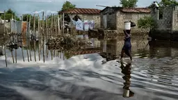 Woman carrying goods on her head running through flooded village. The water is up to her knees and flooded houses can be seen in the background.