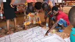 Several people bending town and pointing at a piece of flipchart paper on the floor, with a lot of writing on it.